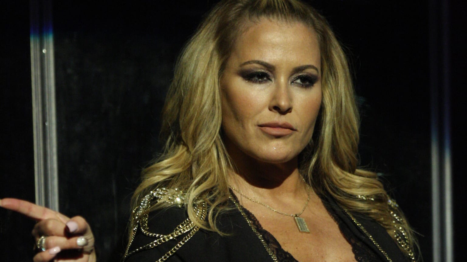 Anastacia on stage in a black jacket with gold chain detail, pointing off to her right