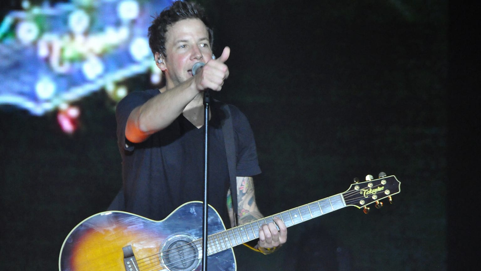 Pierre Bouvier singing into a microphone with an acoustic guitar slung across him, a screen glowing behind
